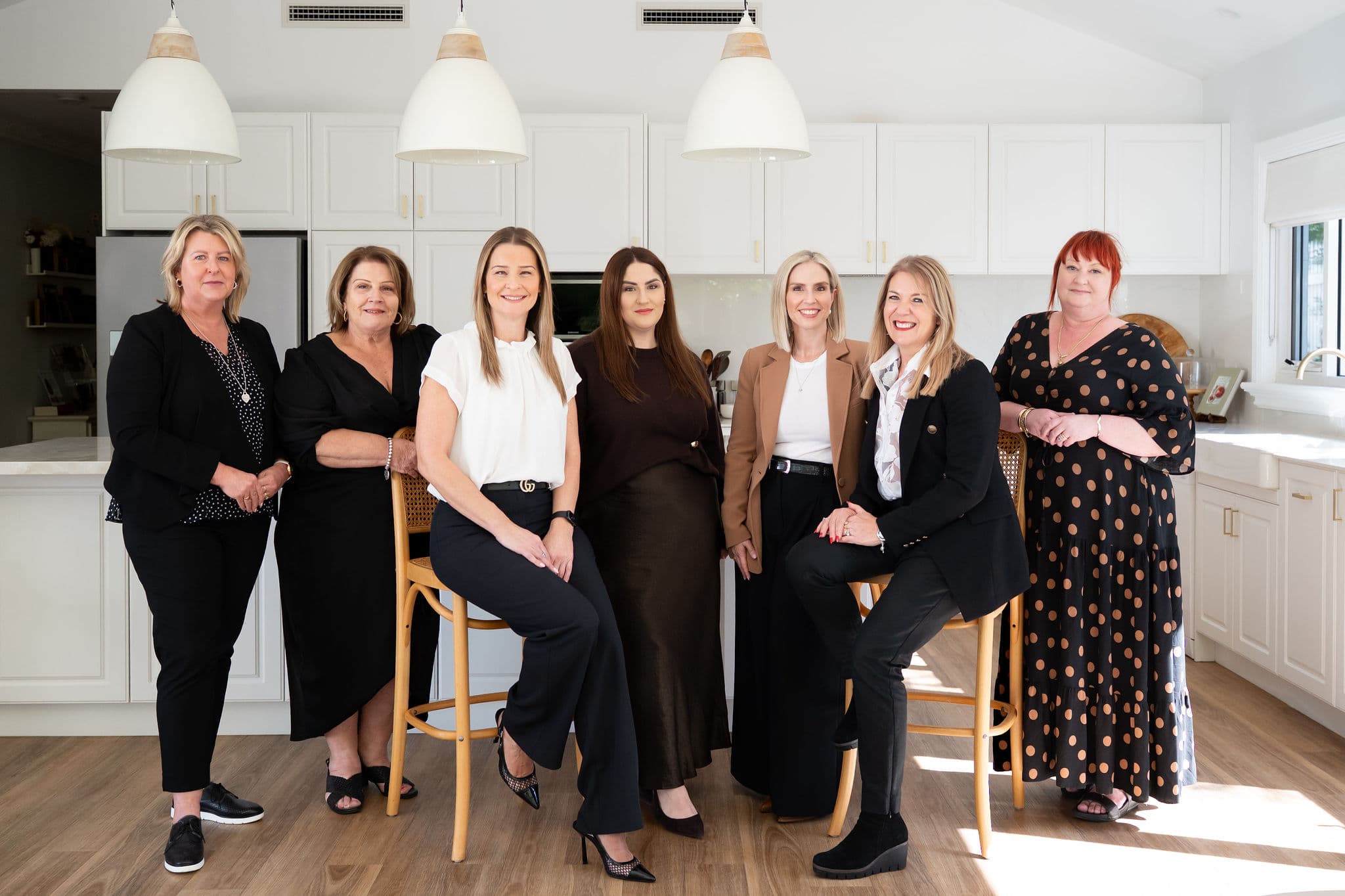 Seven professional women smiling at the camera in a contemporary white kitchen office.