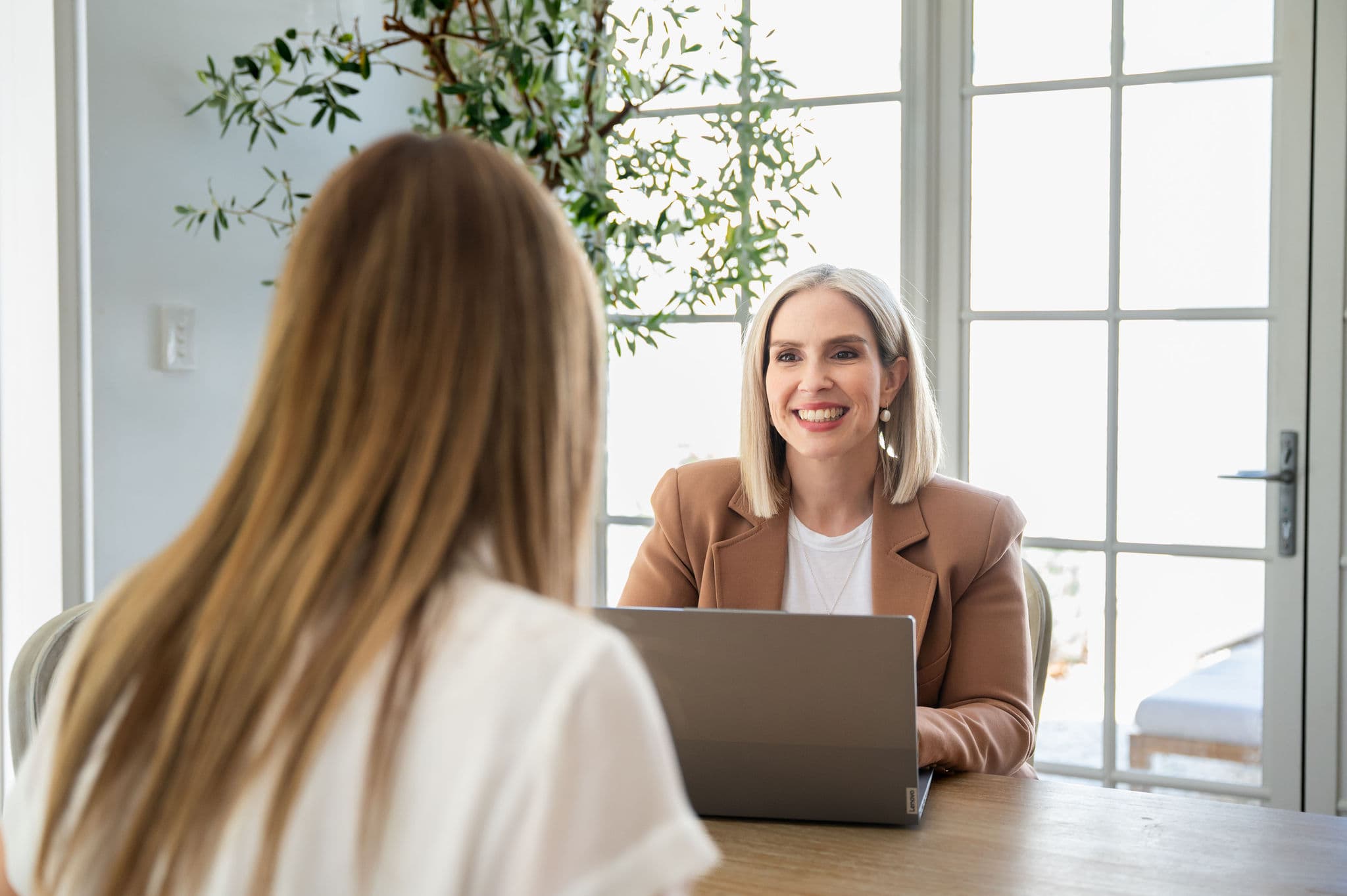 Settlement agent in a tan blazer smiling during a one-on-one client consultation with a laptop on the table.