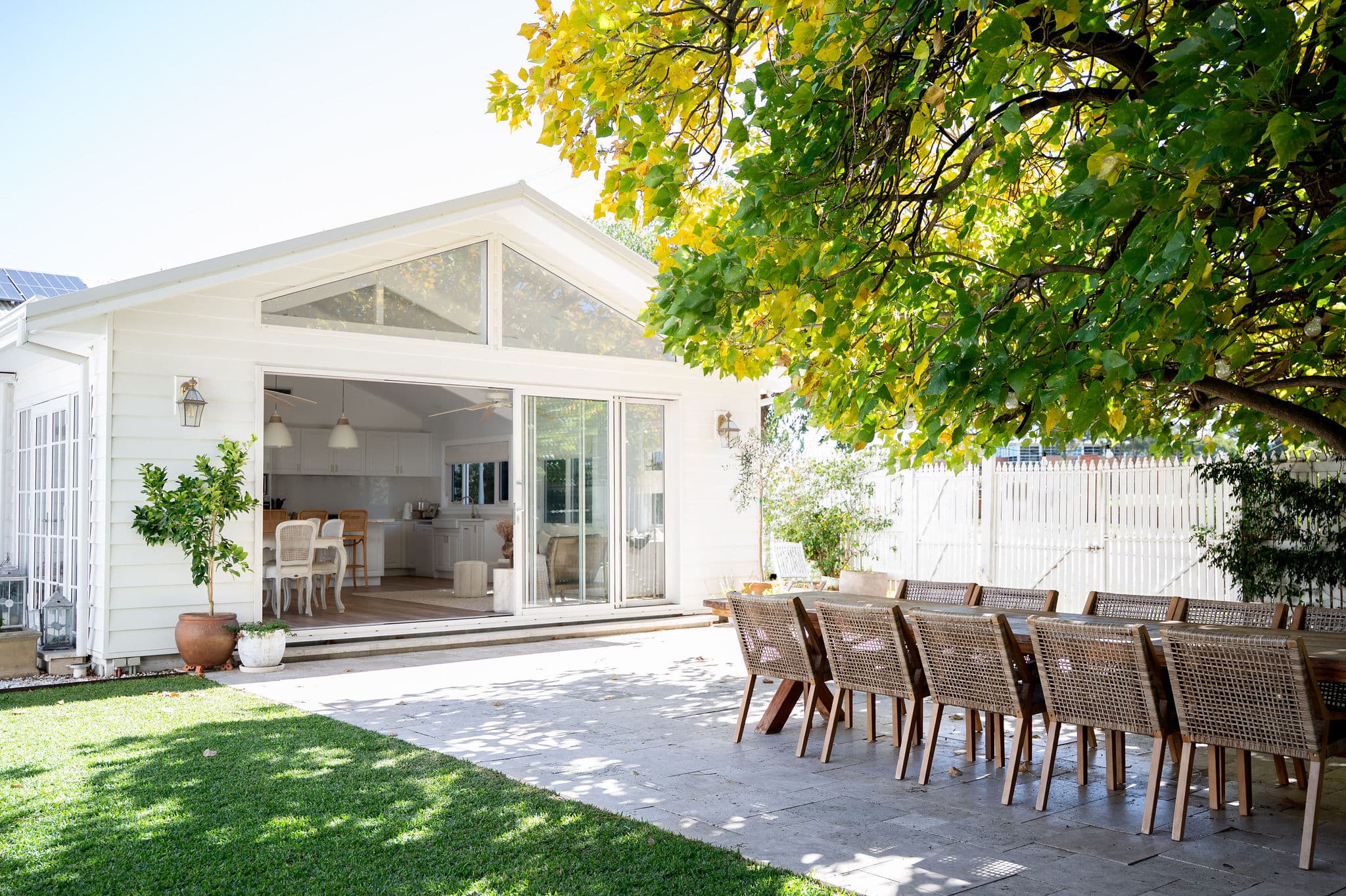 Exterior of a bright white Hamptons-style building opening onto a stone patio with outdoor dining and garden.