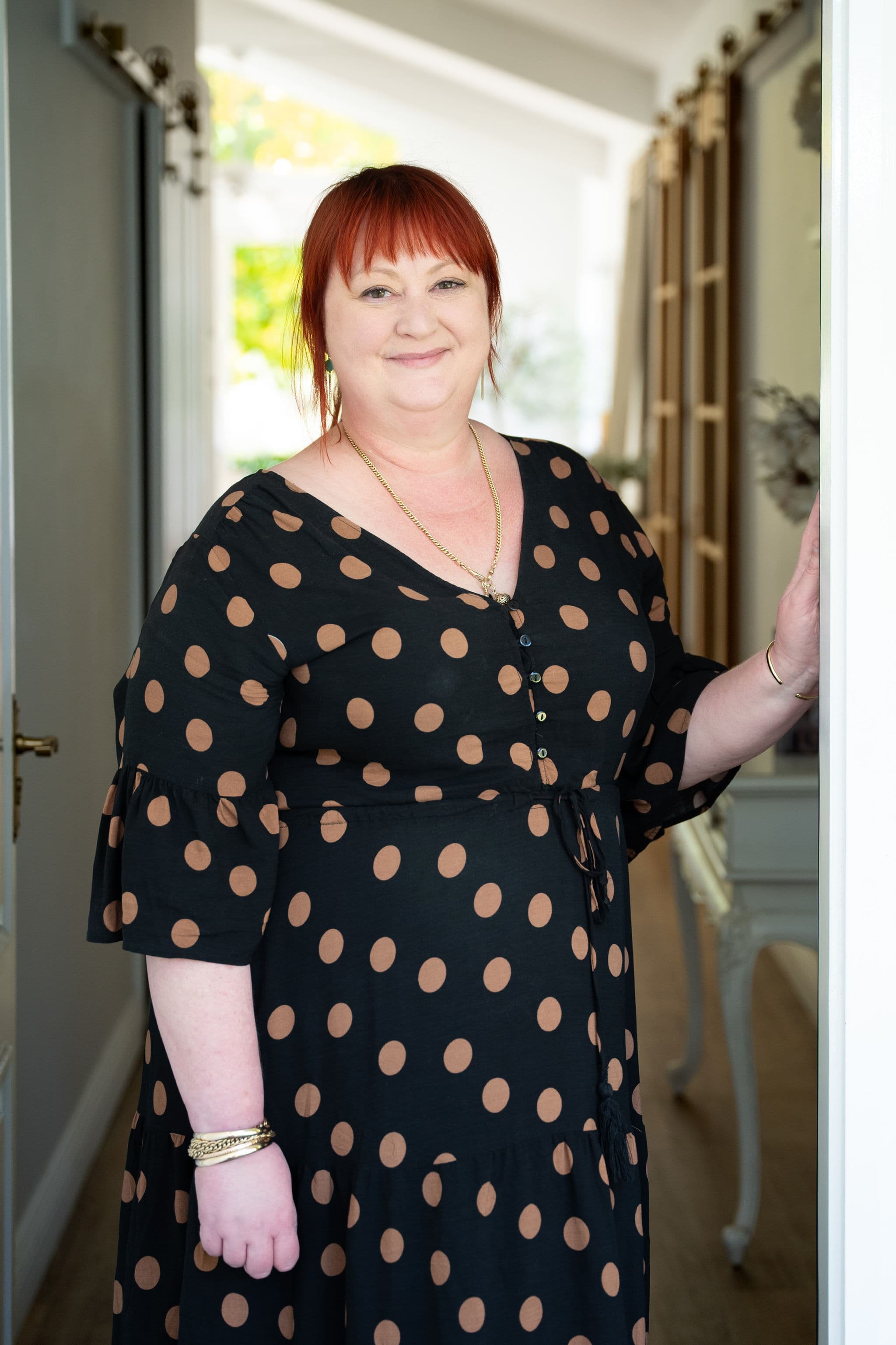 Portrait of Rebecca Cull, conveyancer at West End Settlements, with red hair in a polka-dot dress, standing in a bright doorway.