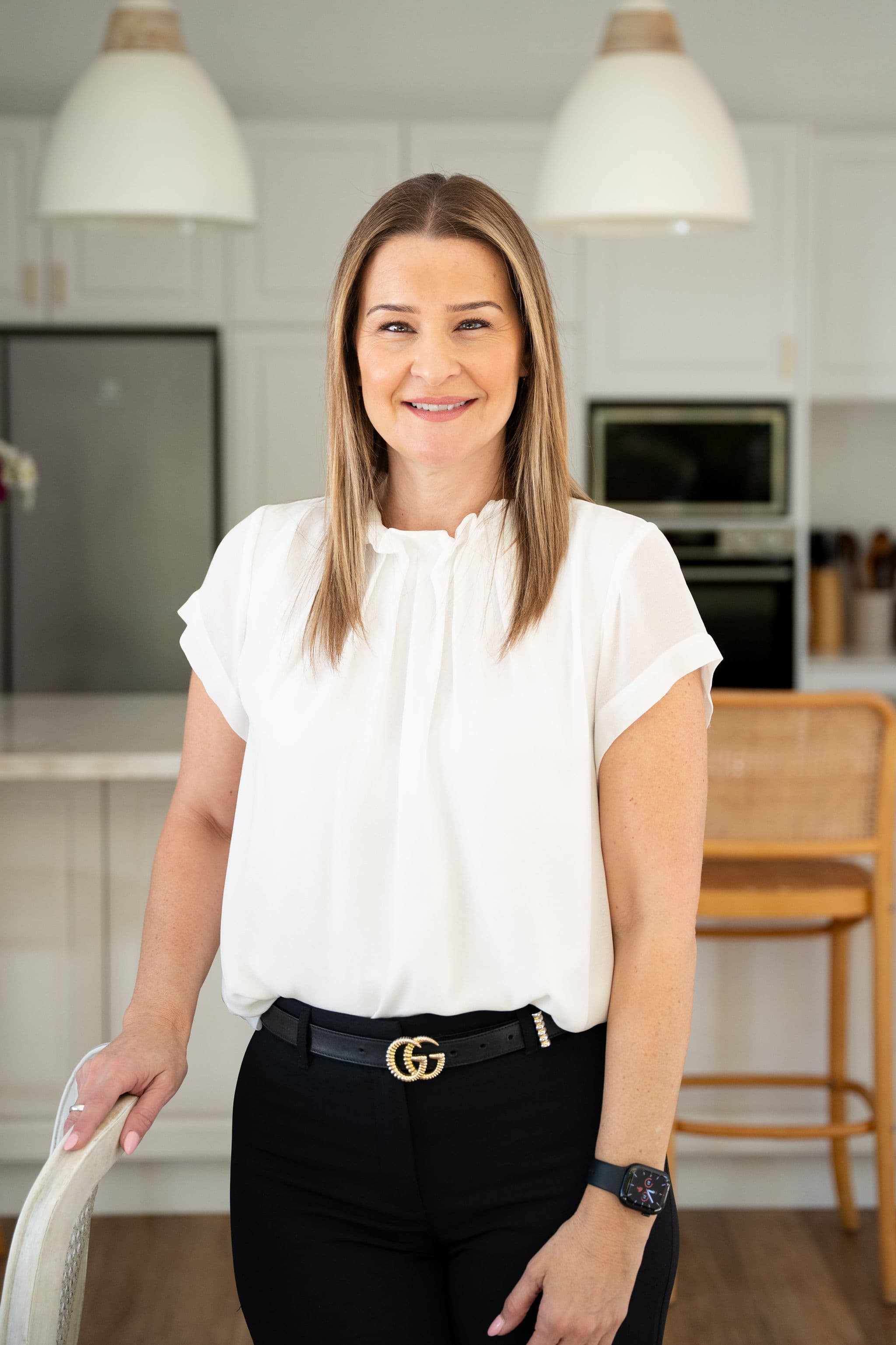 Professional portrait of Zoe Sarris, Settlement Administrator at West End Settlements, in a white blouse smiling in a modern kitchen office.